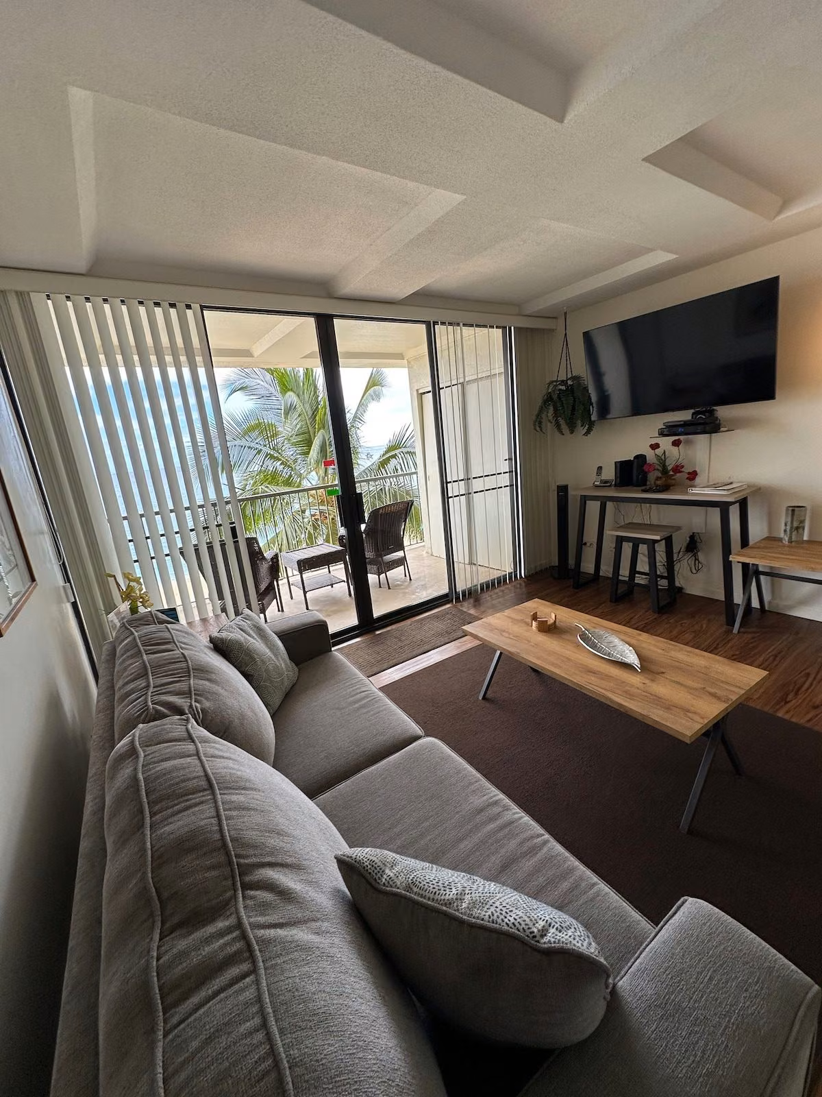 Living room with a grey sectional sofa and wall-mounted TV, looking out through sliding doors to a palm-framed lanai and the ocean